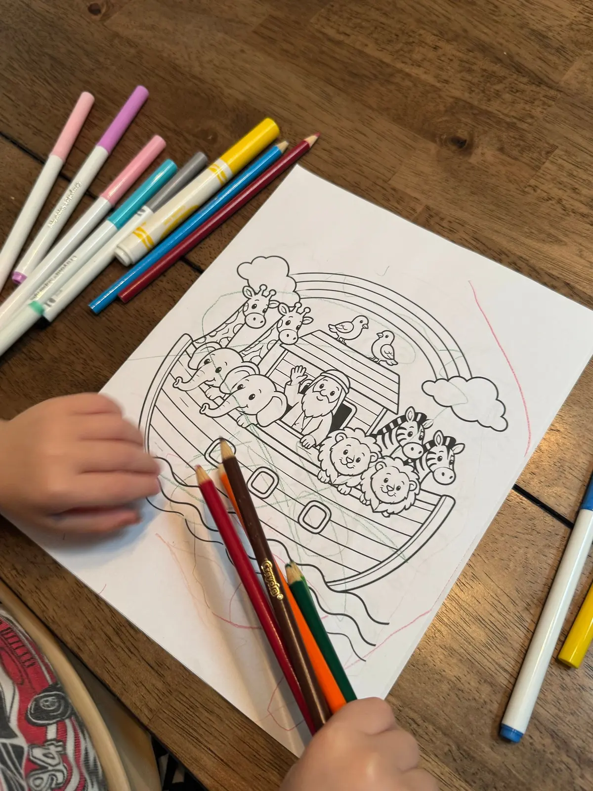 A child's hands coloring a printed Noah's Ark page with colored pencils
