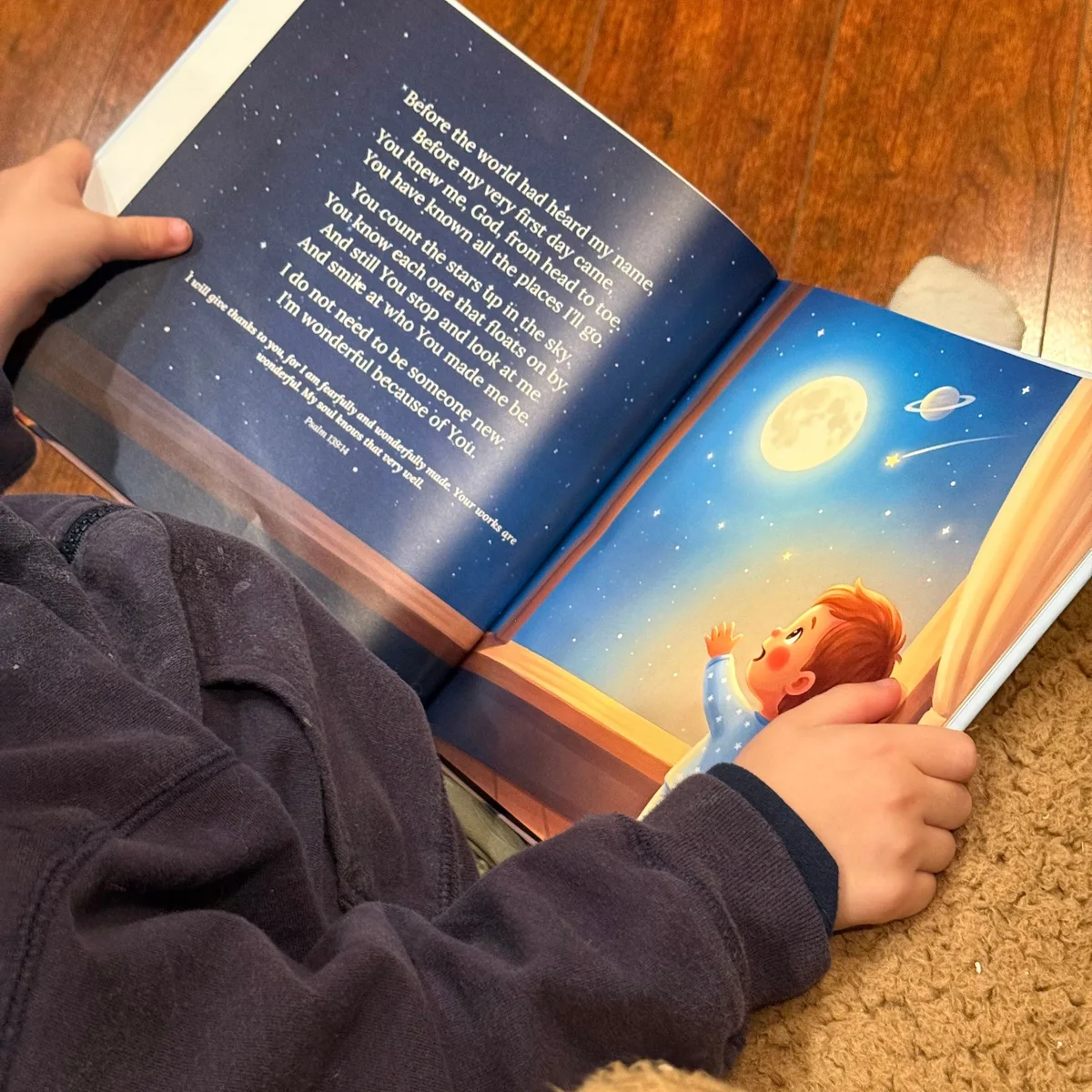 A child's hands holding a printed copy of Little Prayers open at the first prayer, with the moon and stars on the page.