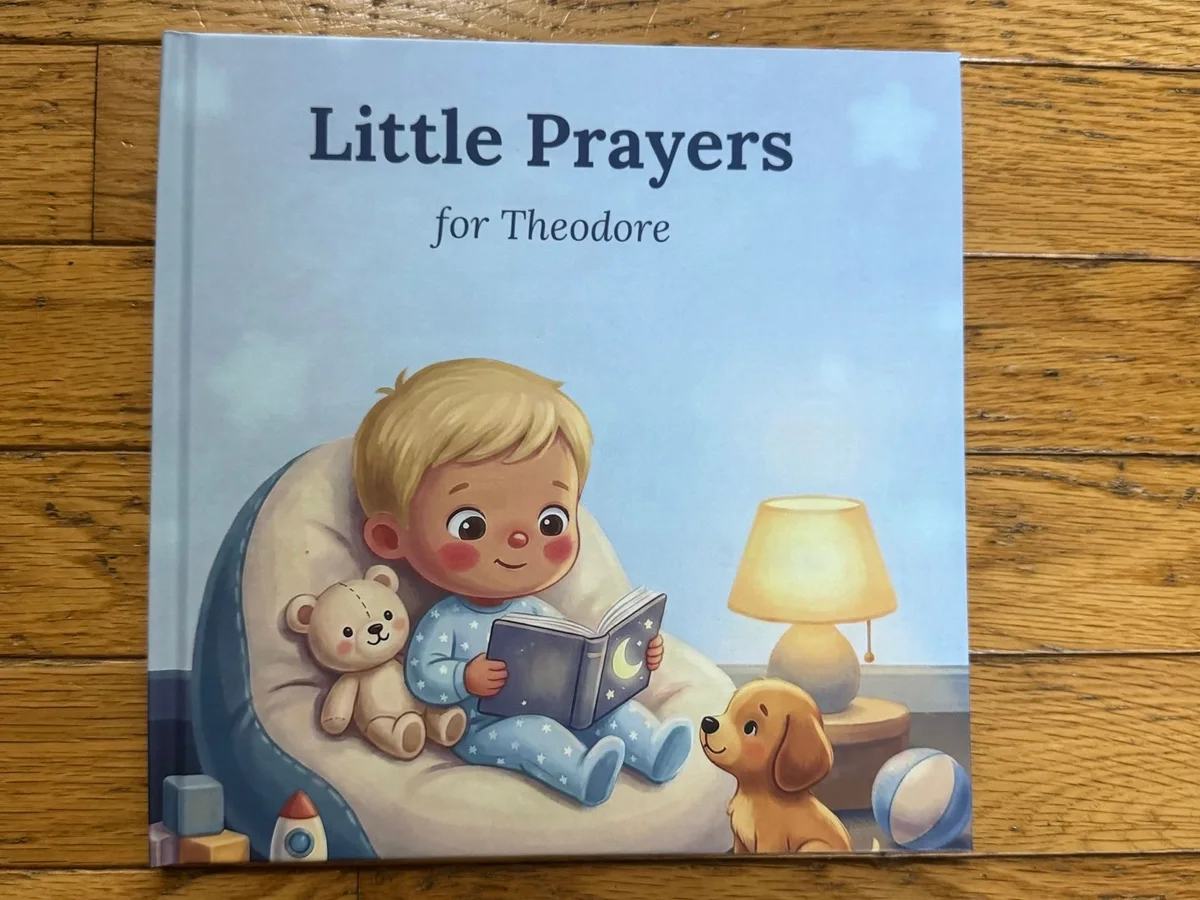 A hardcover copy of Little Prayers lying on a wood floor; the cover reads Little Prayers for Theodore.