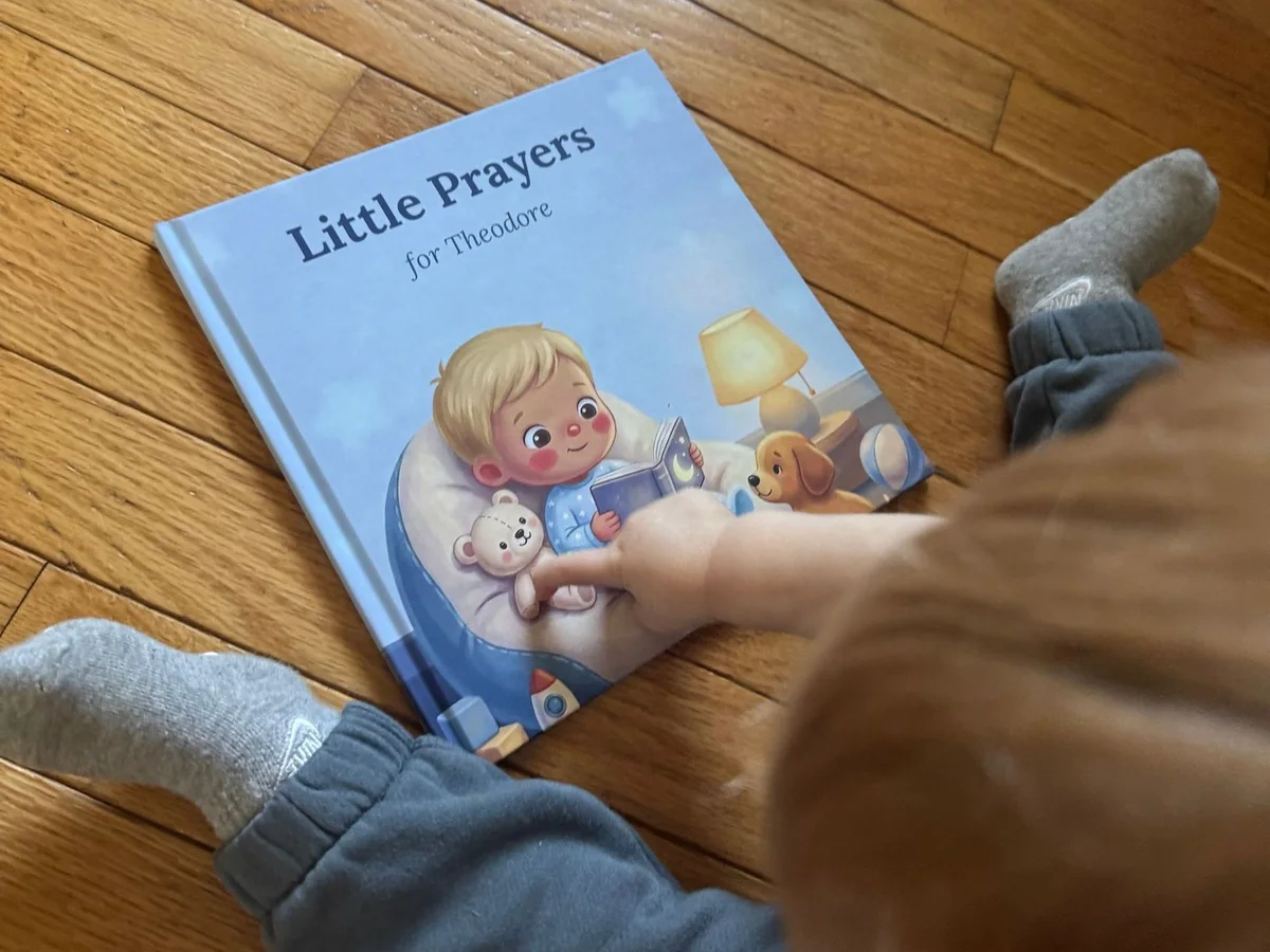 A toddler in grey sweatpants and socks, seen from above, pointing at the cover of a hardcover Little Prayers book on a wood floor.