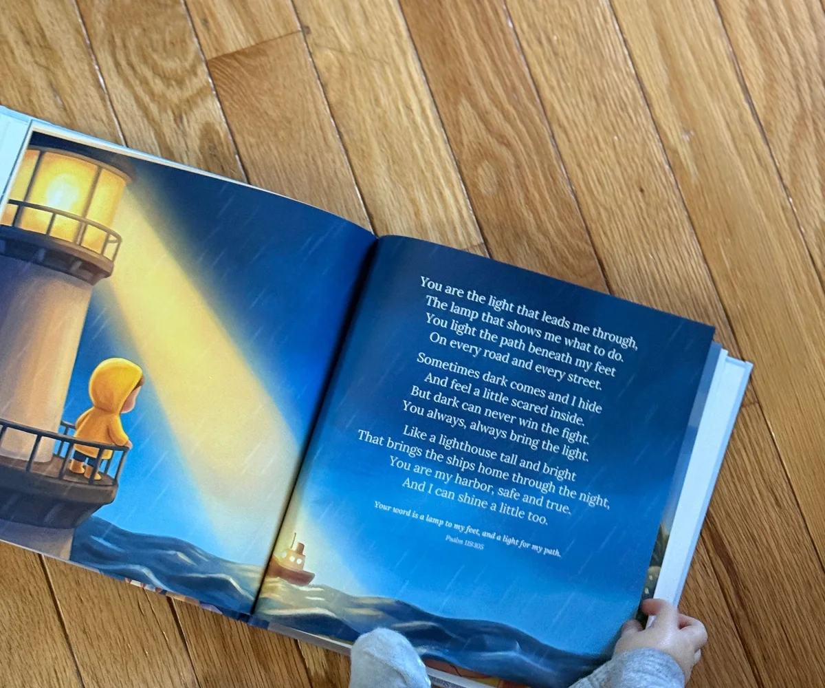 A printed copy of Little Prayers open at the lighthouse spread, lying on a wood floor.