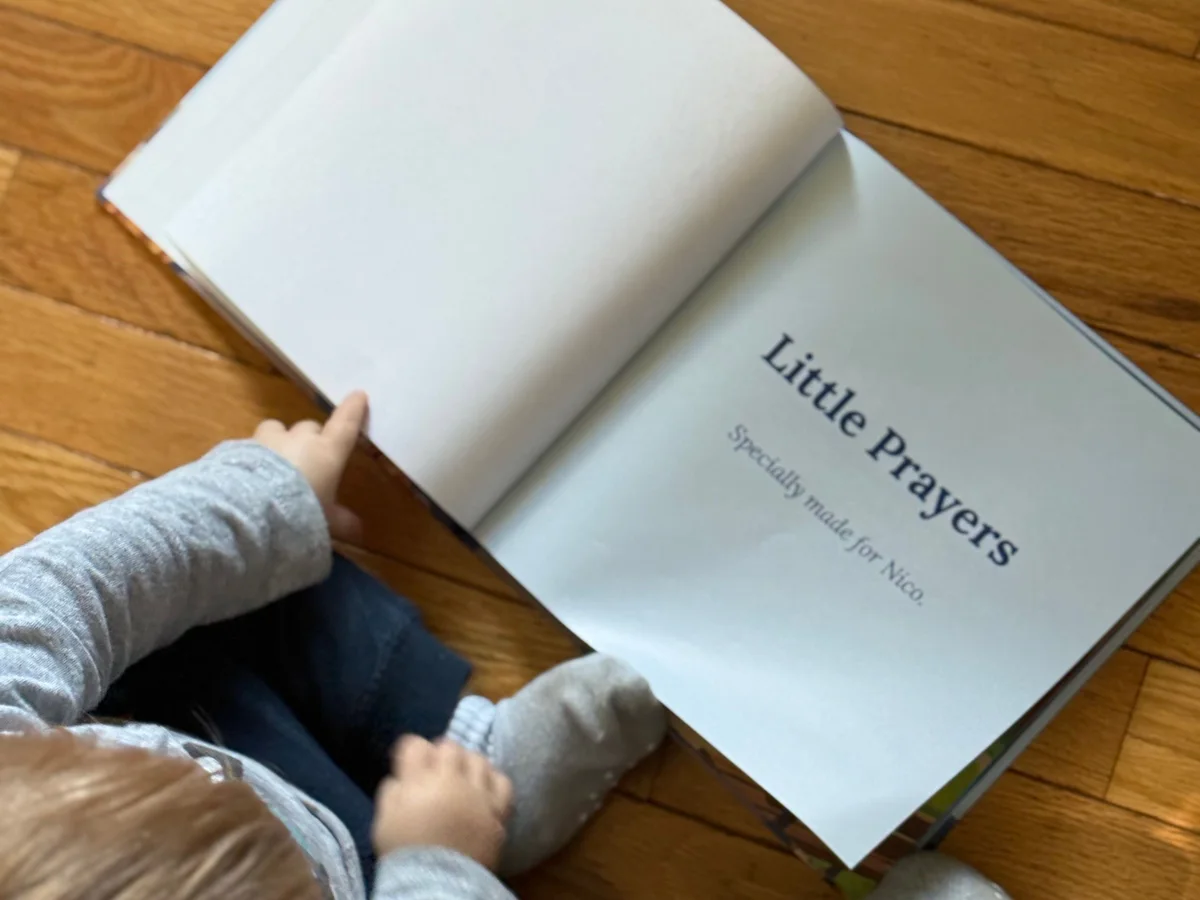 The title page of a printed copy of Little Prayers, open on a wood floor beside a toddler; it reads Specially made for Nico.