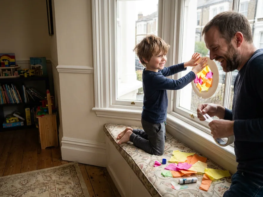 A boy kneeling on a bay window seat pressing a paper-plate sunrise of yellow, orange and pink tissue paper against the glass while his father readies the tape