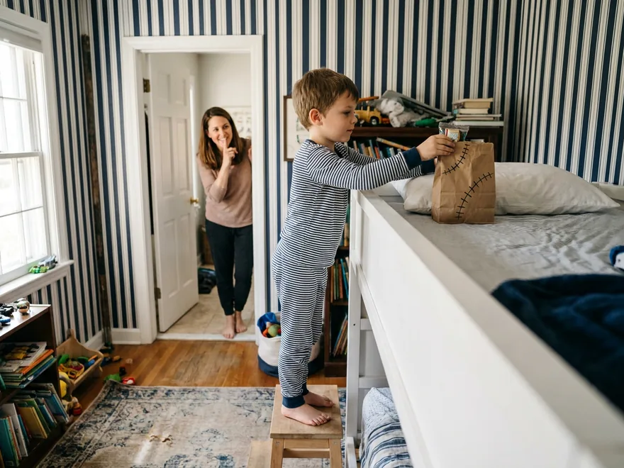 A boy standing on a stool to leave a paper lunch bag drawn with stitches like a grain sack on his brother's top-bunk pillow while his mother watches from the doorway