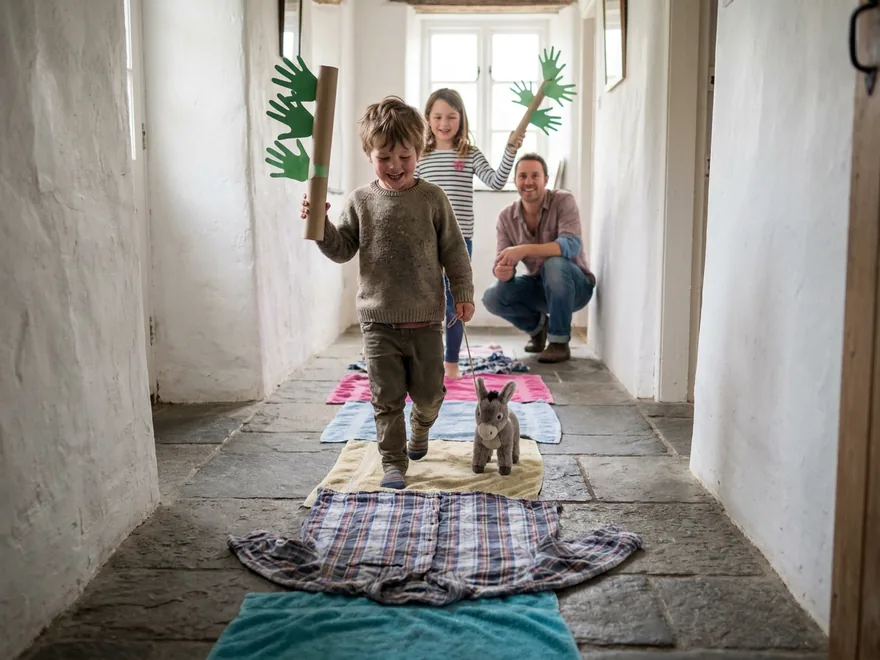 A boy parading down a stone-floored cottage corridor over a line of towels and old shirts, waving a paper handprint palm branch and pulling a toy donkey, his sister and father behind