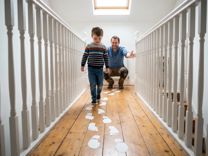 A boy walking slowly along a winding line of paper footprints on an upstairs landing toward his father, who crouches at the far end reaching out to him